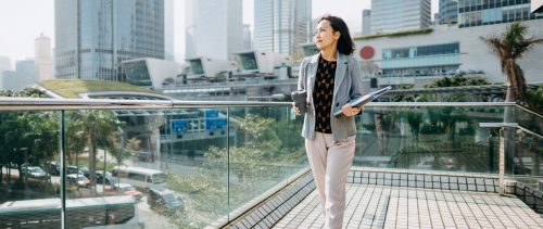 Portrait of confidence Asian businesswoman looking away and holding documents and having coffee to go against city scene in front of modern office buildings