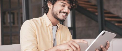 Happy Indian man shopping online, ordering food, reading good news sitting on sofa. Smiling asian freelancer using digital tablet, receive payment working online from home